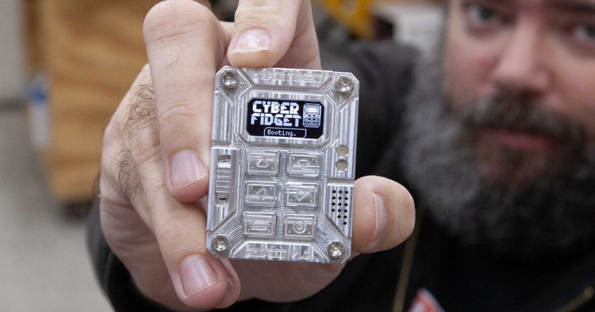 A hand holding the machined-aluminum Cyber Fidget, showing its screen, buttons and slider